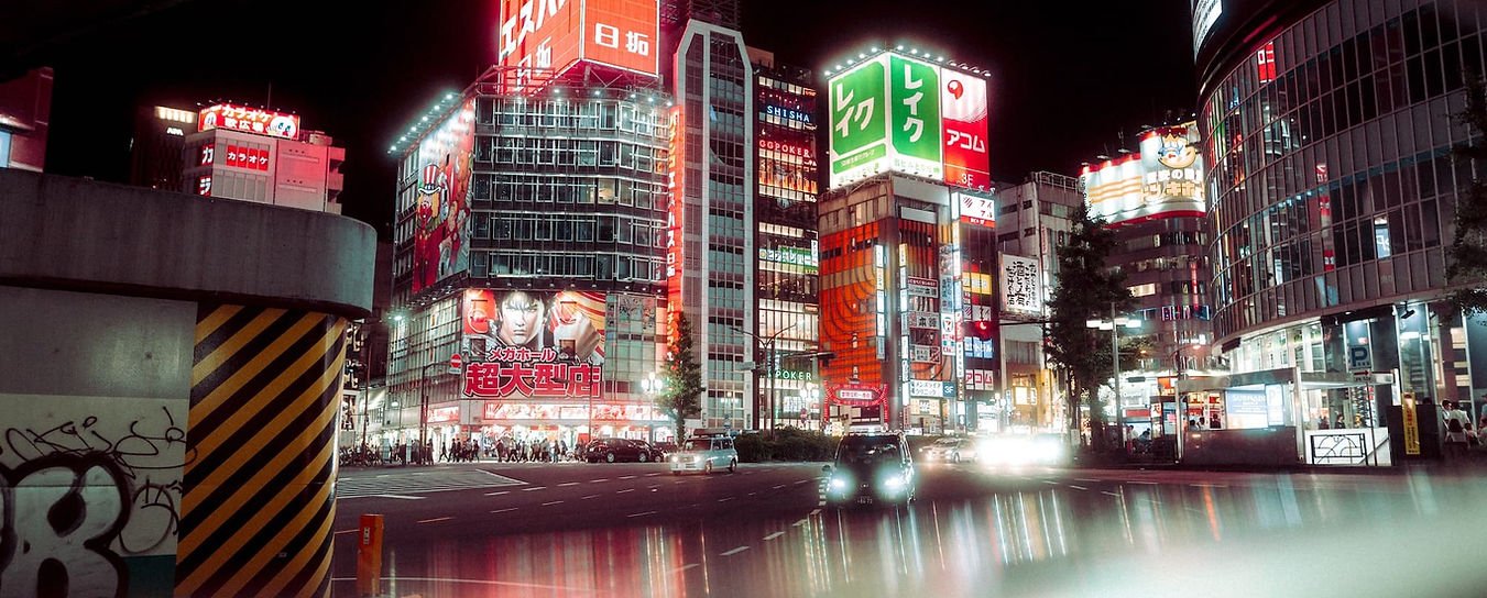 Nighttime city street in Japan with illuminated billboards, neon signs, and cars. Modern buildings and pedestrians are visible.