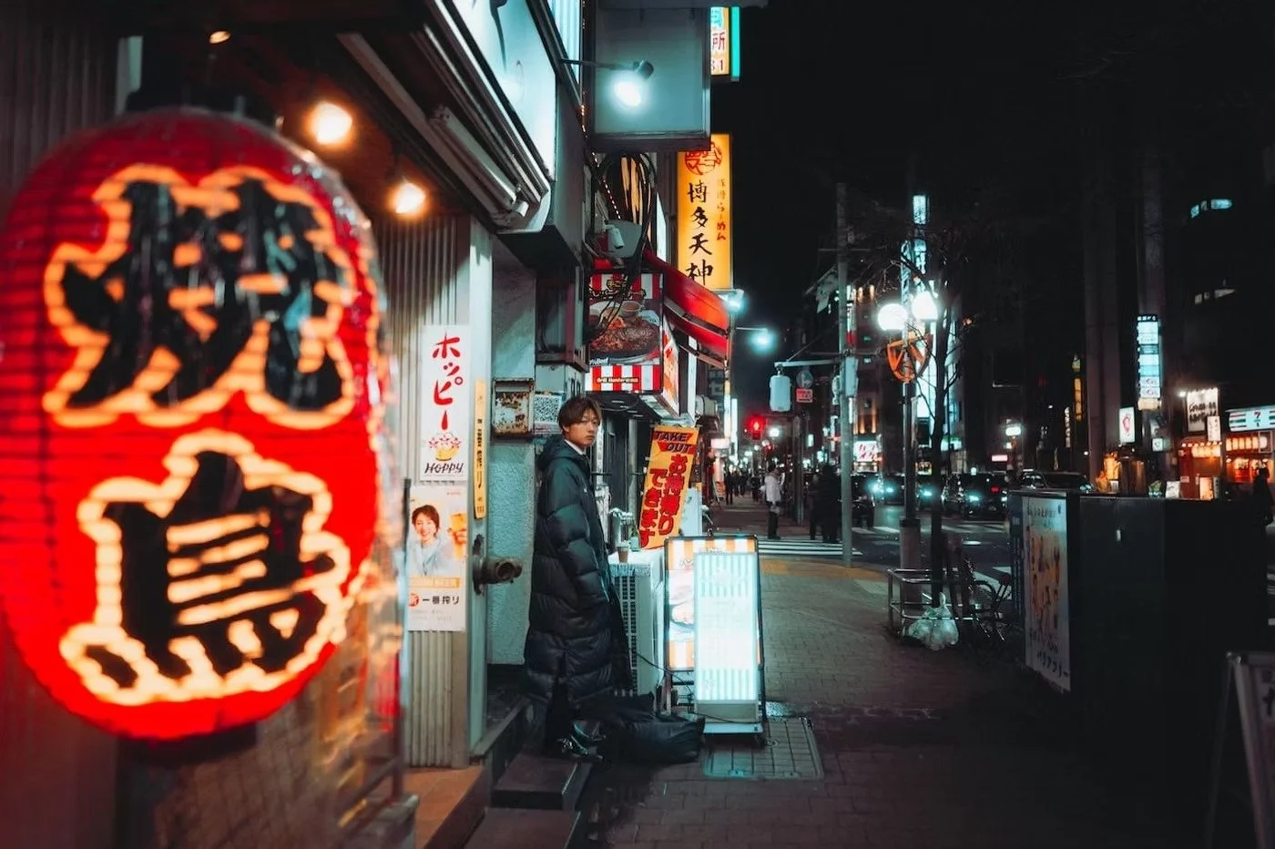 Nighttime street scene in Japan with neon signs, a man in a puffy coat next to a small restaurant, and a red paper lantern in the foreground. Shimbashi night scenes on the Tokyo Afterhours street photography tour.