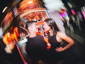 A man and woman talking and smiling at each other in a lively, colorful indoor setting with blurred lights.