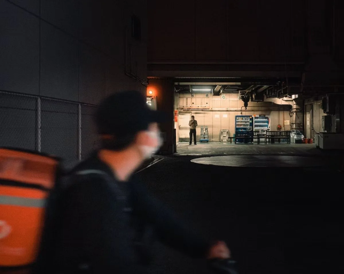 A person in the foreground wearing a face mask and backpack, looking toward a person standing in a brightly lit garage or loading area in the background.