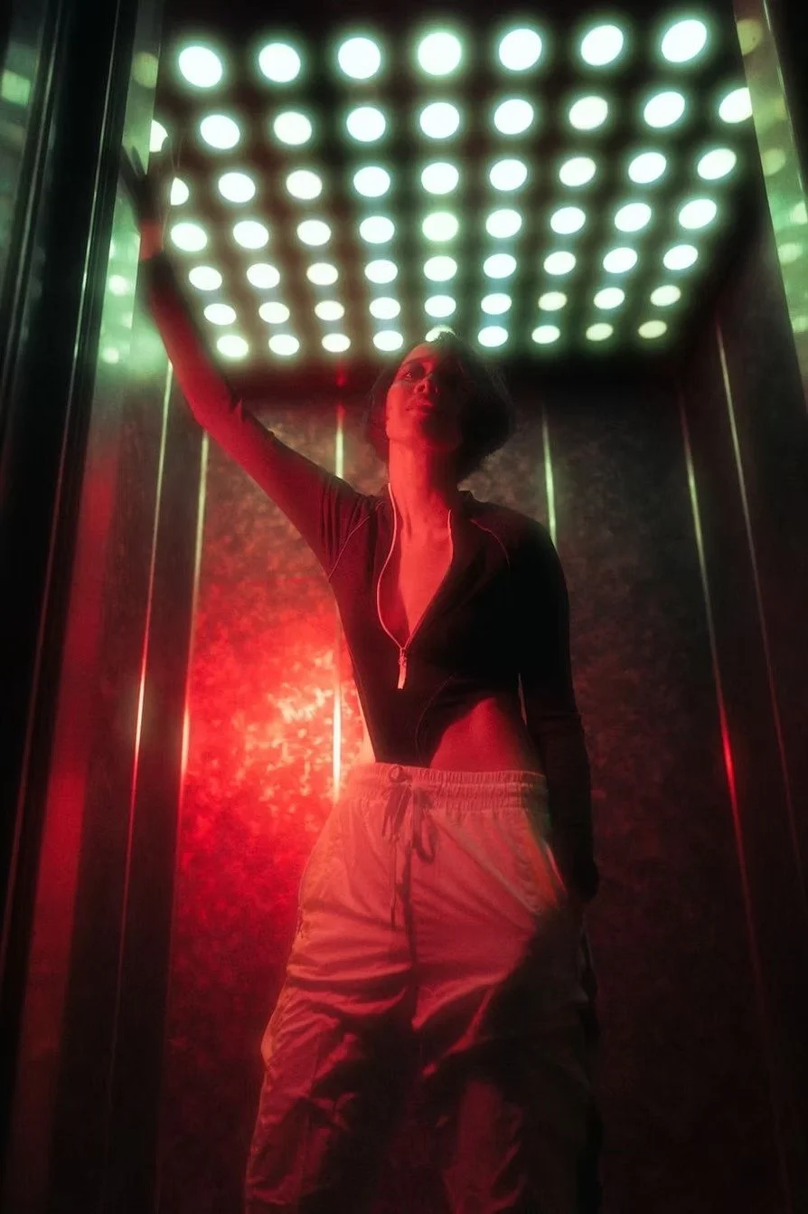A woman inside a small, enclosed space with a ceiling of bright white LED lights. She is posing with her left arm raised, wearing a black zip-up top and beige cargo pants, illuminated by red and white lighting shot by portrait photographer Deniz Demi