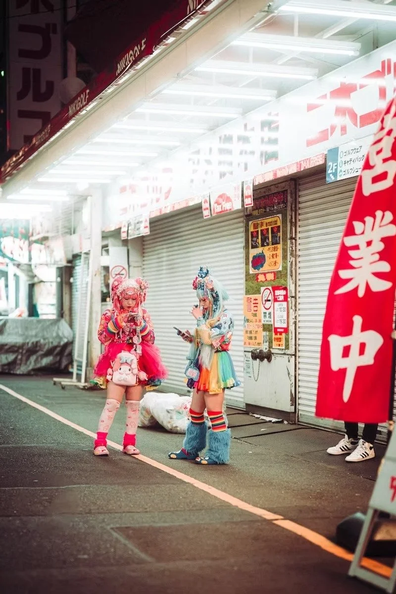 Two people dressed in colorful, elaborate costumes with pastel and rainbow hues, standing on a street at night. They are looking at their phones in front of closed storefronts.