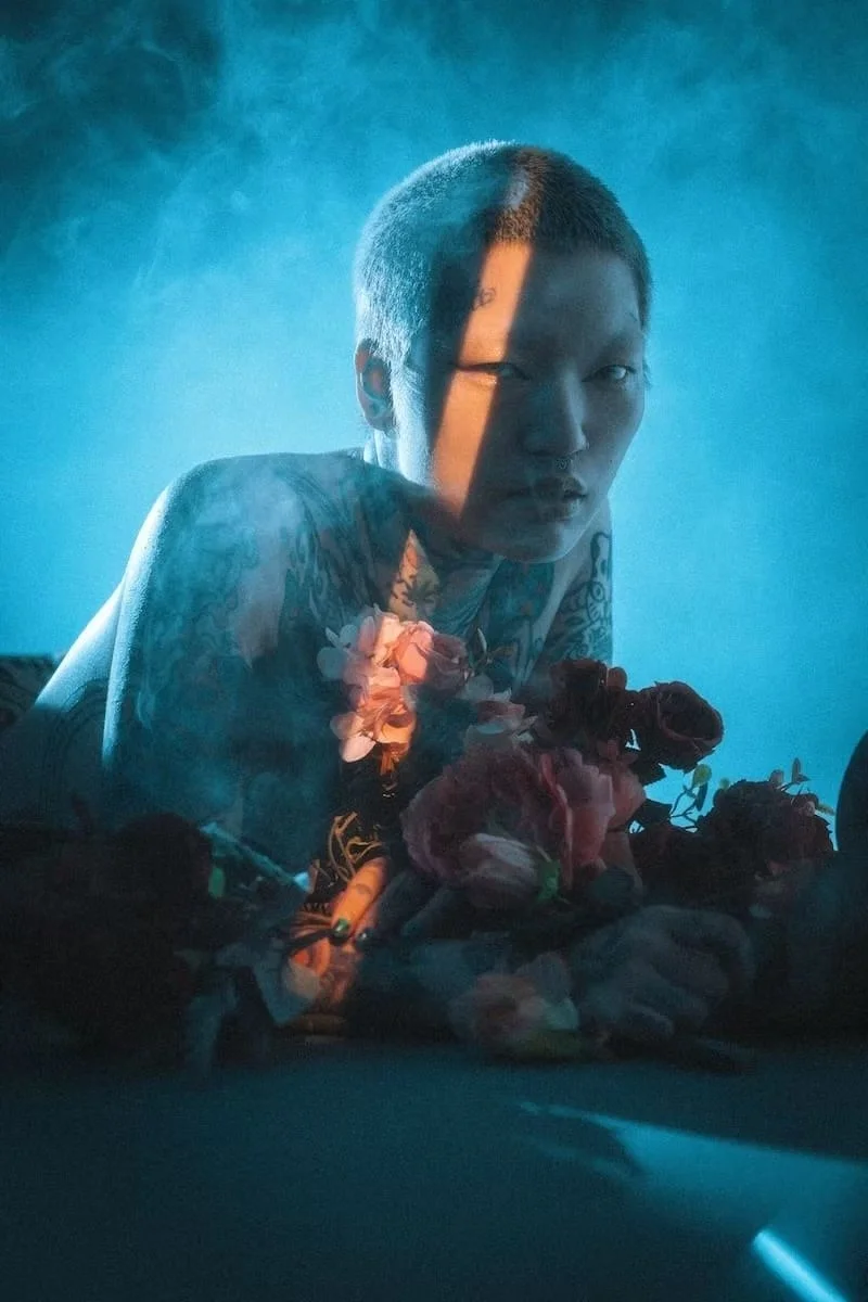 Person with short hair and tattoos resting their arm on a surface with flowers, illuminated by blue light with a shadow cast across their face.