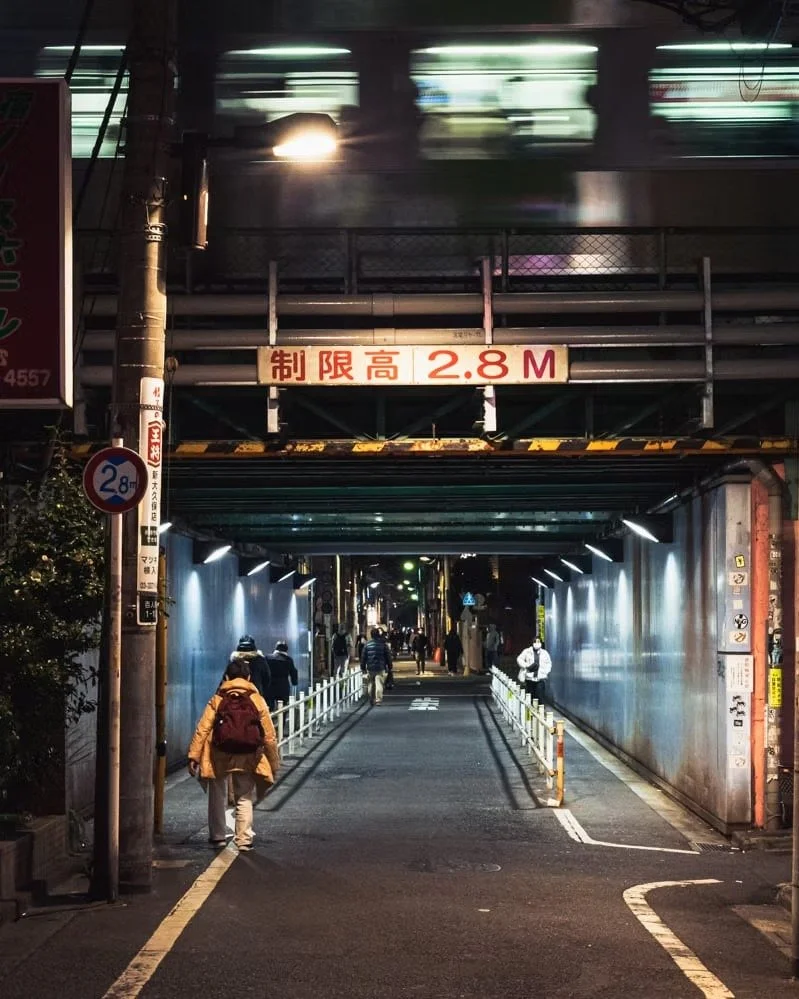 Night scene of a city street under a bridge or overpass with pedestrians walking and a blurred train passing overhead. Sign indicates a height limit of 2.8 meters.
