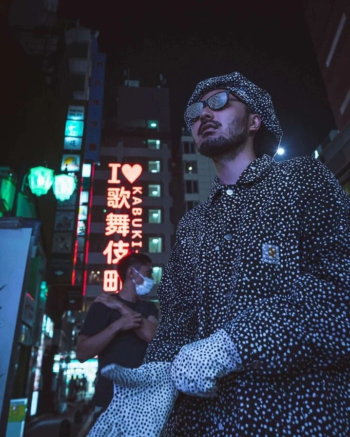 A man in a polka-dot outfit and sunglasses standing under neon lights, with a "I ❤️ Kabukicho" sign behind him.