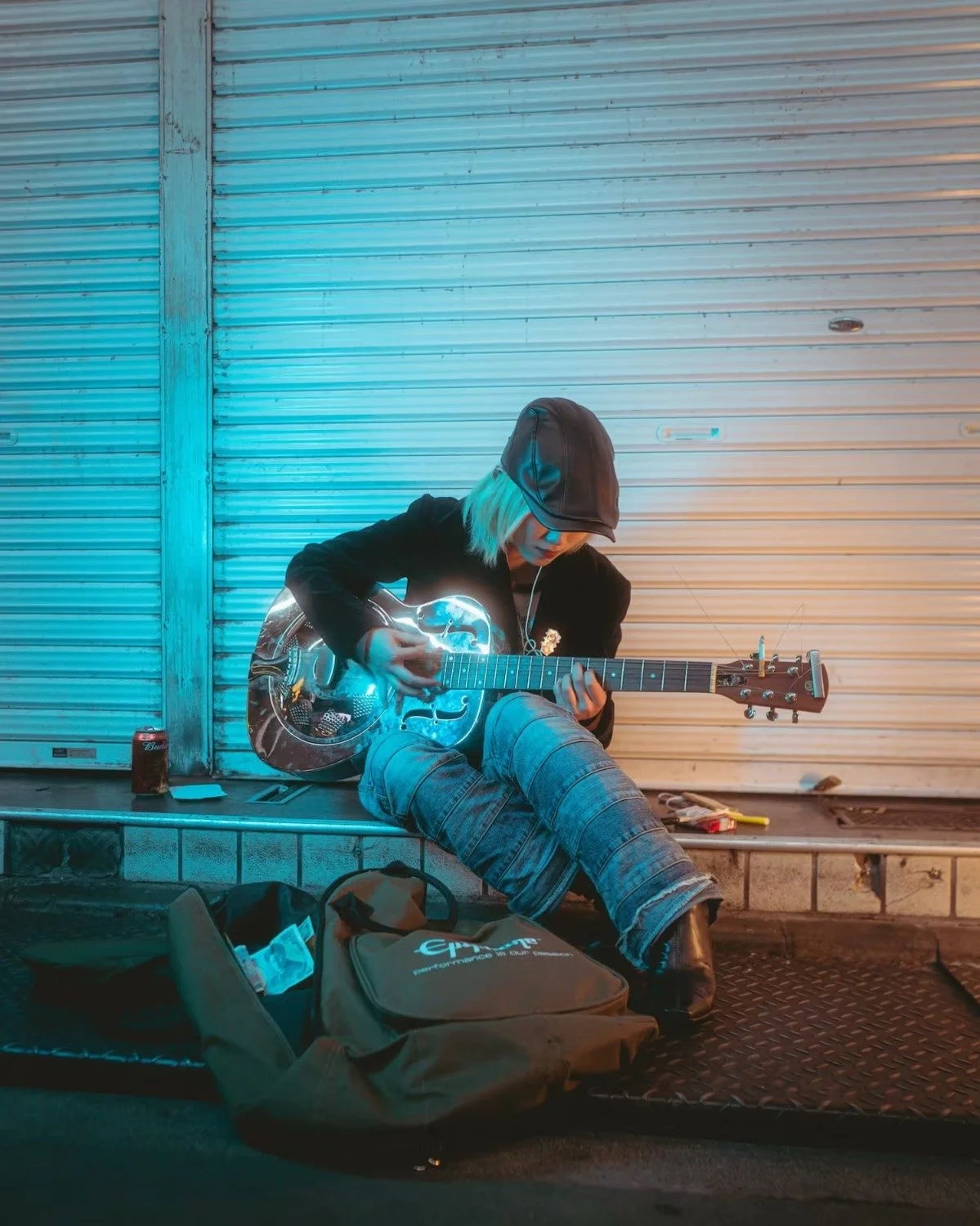 A person with long blonde hair, wearing a black leather cap and jacket, sitting on a step in front of a metal roll-up door, playing an electric guitar illuminated with neon-like lighting, with a brown bag and drink can nearby in Koenji Tokyo.