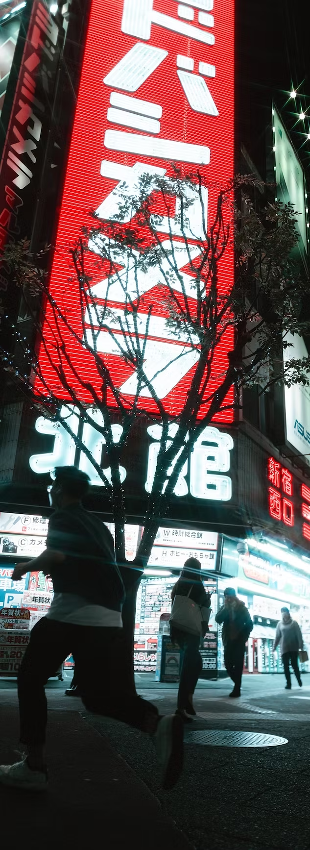 Bright neon signs in Japanese on a city street at night, with silhouettes of people walking in front of a small tree.