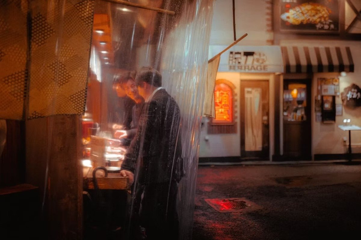Two men in suits standing close together and looking at their phones outside a restaurant or store at night, with warm lighting and some signs and flowers on the building front. Nakano at night on the Tokyo Izakayas Street photography Tour.