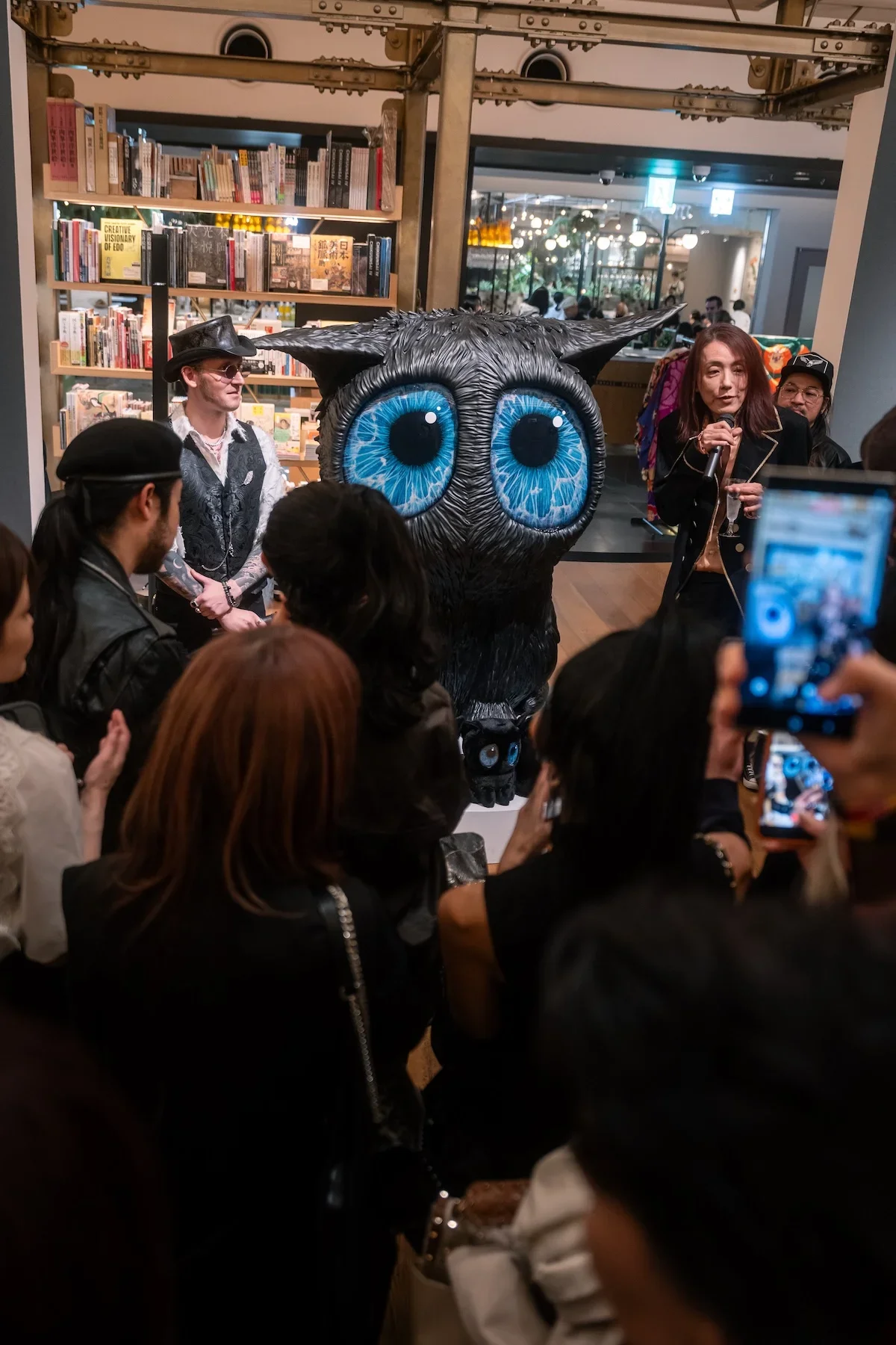 August Vilella
---
Visitors gather around a large, dark sculptural creature with striking blue eyes during August Vilella's exhibition, documenting the immersive artwork with smartphones and cameras.