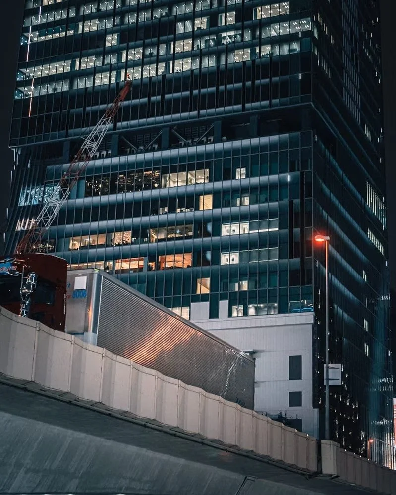 Night view of a city high-rise building with illuminated windows, a construction crane, and part of a highway with a barrier in the foreground.