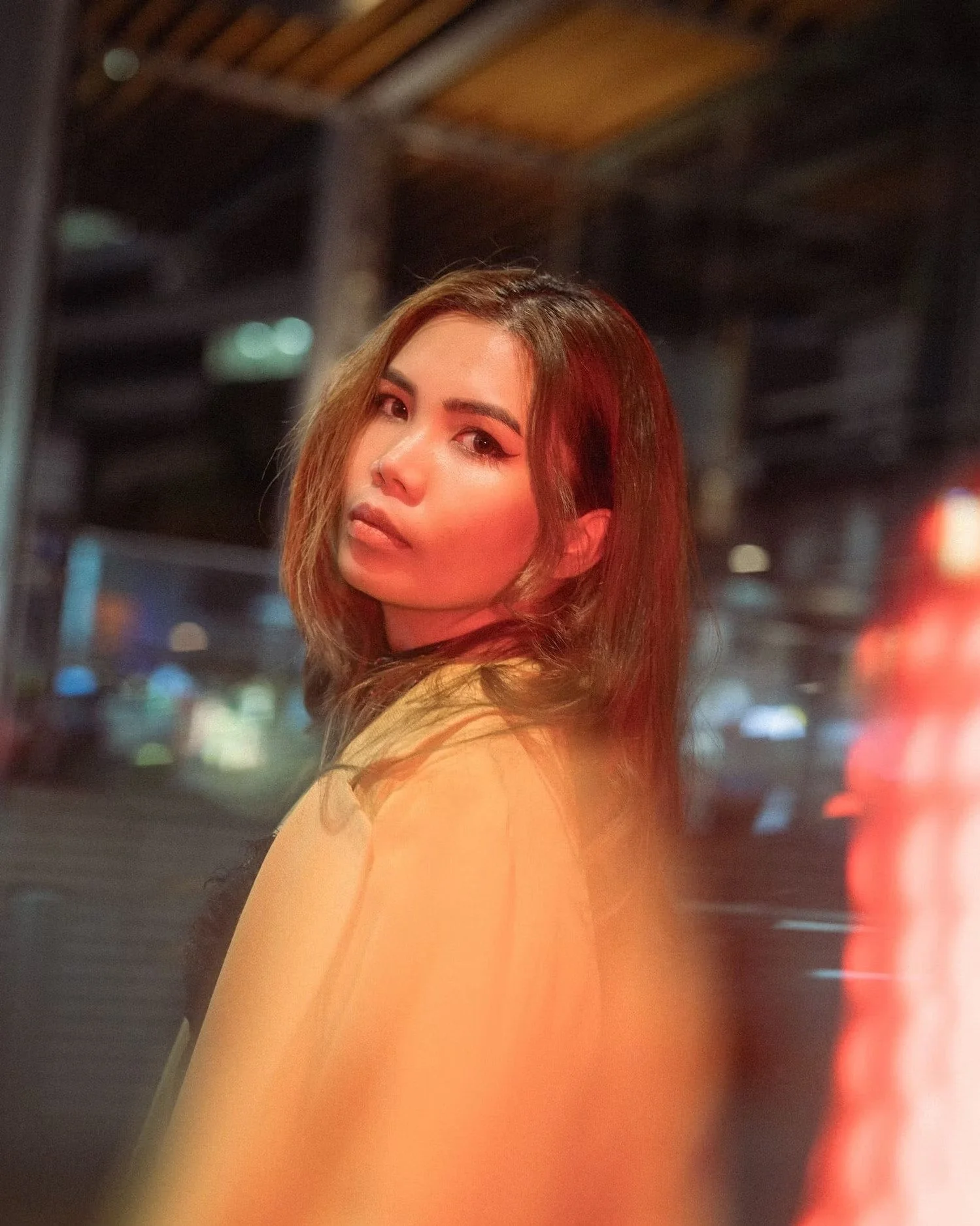 A young woman with long, wavy brown hair looking over her shoulder at the camera in a dimly lit urban setting at night with colorful bokeh lights.