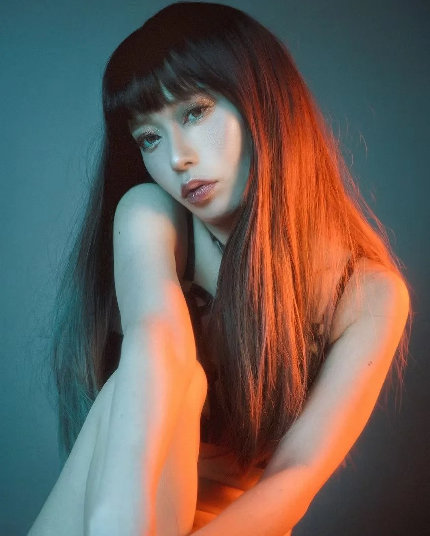 A model with long flowing hair stands gracefully in a soft blue-lit environment. The moody lighting highlights her posture and creates a dreamy, ethereal effect, making her the focal point of the image.