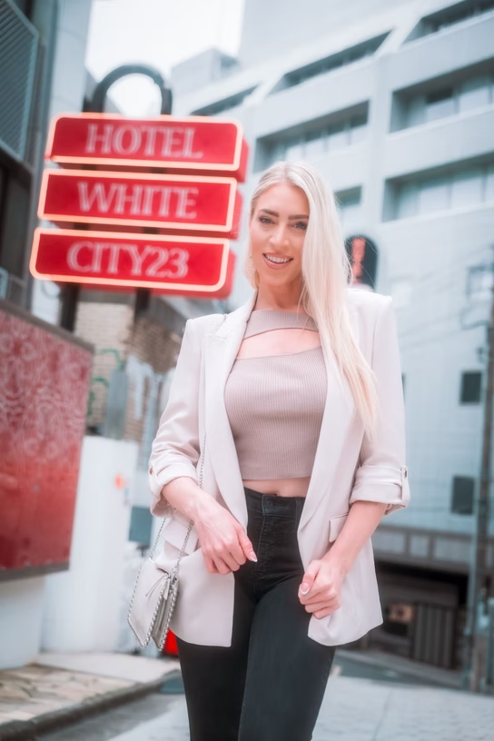 A young woman with long blonde hair smiling and walking outside in an urban area, with a red lit hotel sign that reads 'HOTEL WHITE CITY23' in the background.