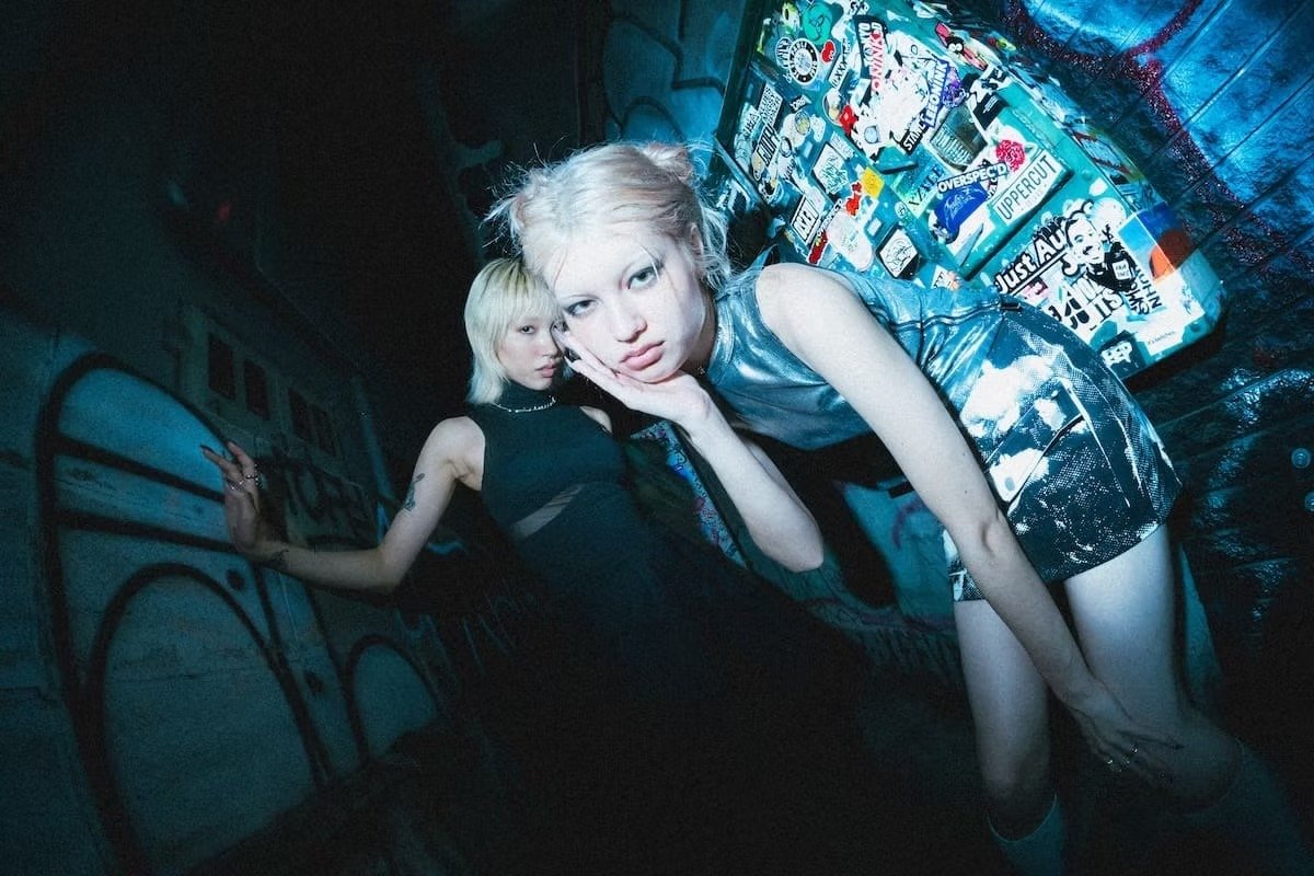 Two models pose in a narrow Tokyo alley at night, lit with cool blue flash and neon spill, wearing metallic and black Fragile Base garments. One leans toward the lens while another stands behind against graffiti-covered walls, creating a raw, undergr