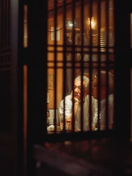 A person sitting at a table seen through a wooden lattice or partition in a dimly lit room.