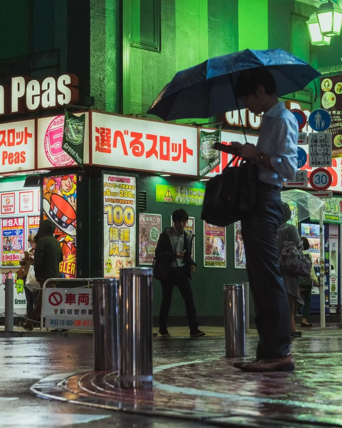 A man holding a blue umbrella and using a smartphone standing on a rain-soaked street at night, with a busy neon-lit storefront and other pedestrians in the background. Shot by Deniz Demir.