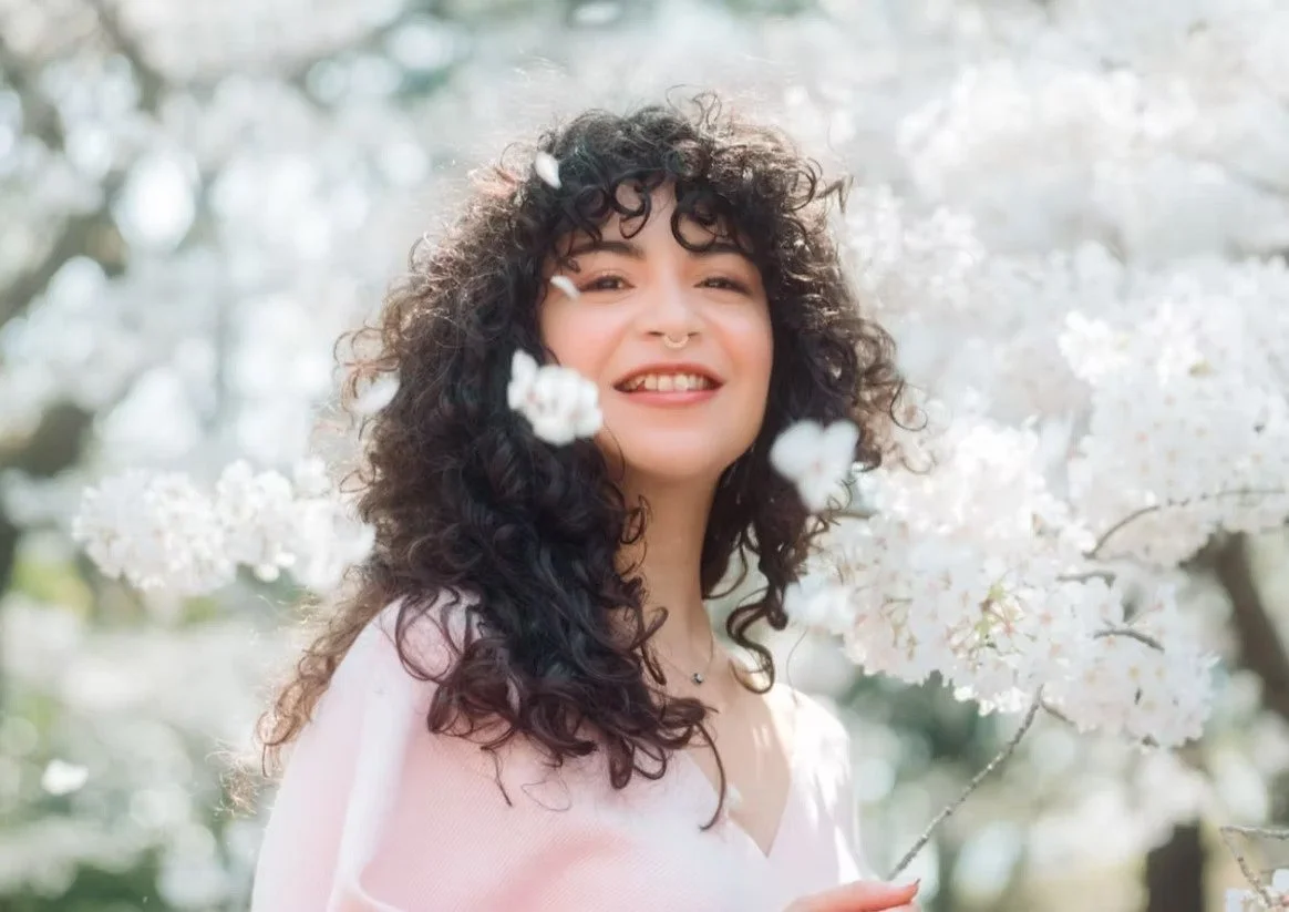 A woman with curly hair smiles at the camera, surrounded by cherry blossoms. The soft petals float around her, creating a dreamy and joyful springtime scene.