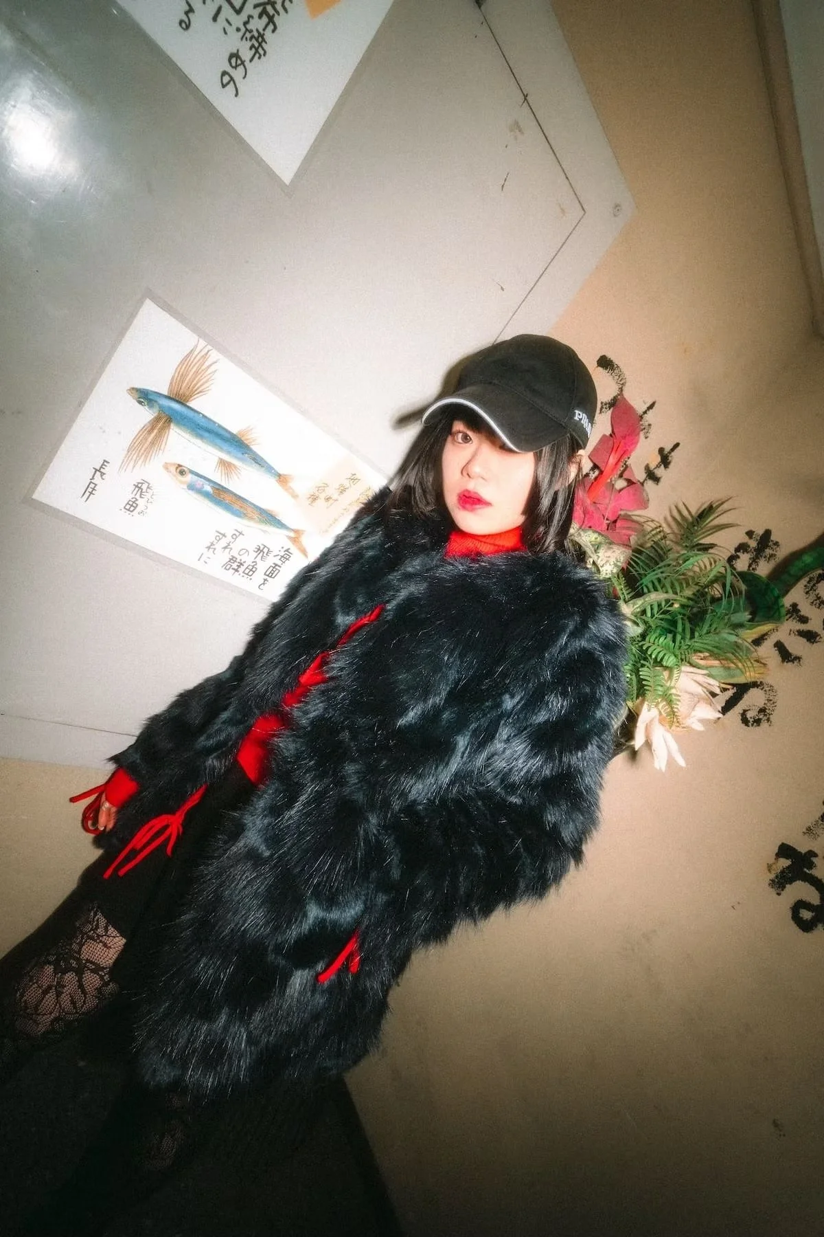 A woman with black hair and red lipstick wearing a black cap and a black faux fur coat with red accents stands indoors near Japanese fish posters and a plant with red and white flowers.