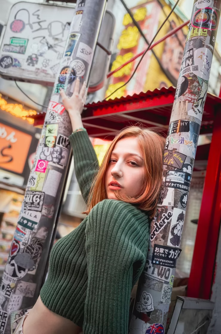 A young woman with reddish-brown hair, wearing a green ribbed sweater, leaning against two sticker-covered poles in an urban setting with graffiti and colorful signs in the background.