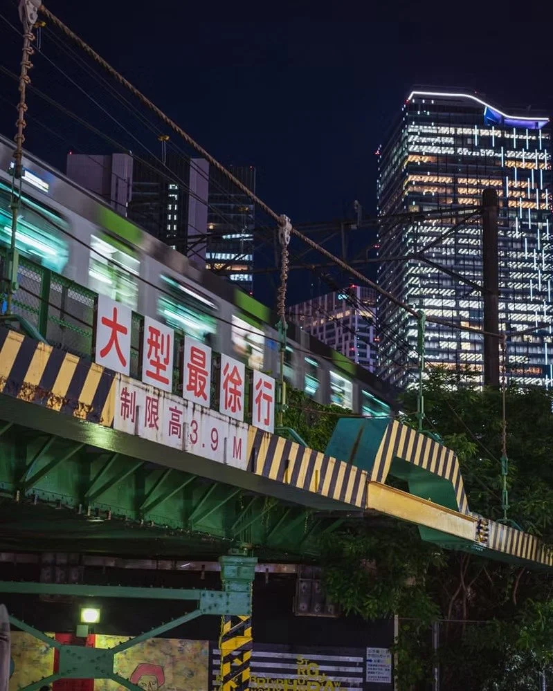 A light rail train passing over a bridge at night in an urban area, with tall illuminated modern buildings in the background. A sign on the bridge has Japanese characters and indicates a height limit of 3.9 meters. Shibuya.