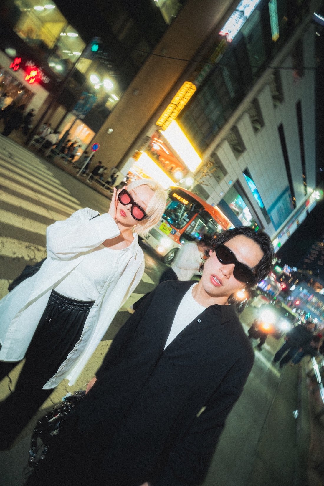Vasuma Eyewear
---
Two stylish young adults, one with blonde hair and pink sunglasses and the other with dark hair and black sunglasses, standing on a busy city street at night with illuminated signs, buses, and pedestrians. Shot for Vasuma Eyewear b