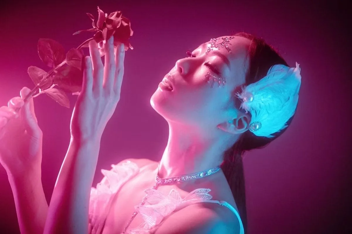 A woman with closed eyes holding a flower, illuminated by pink and blue lights, wearing pearl earrings, a feather accessory in her hair, and sparkling makeup on her face.