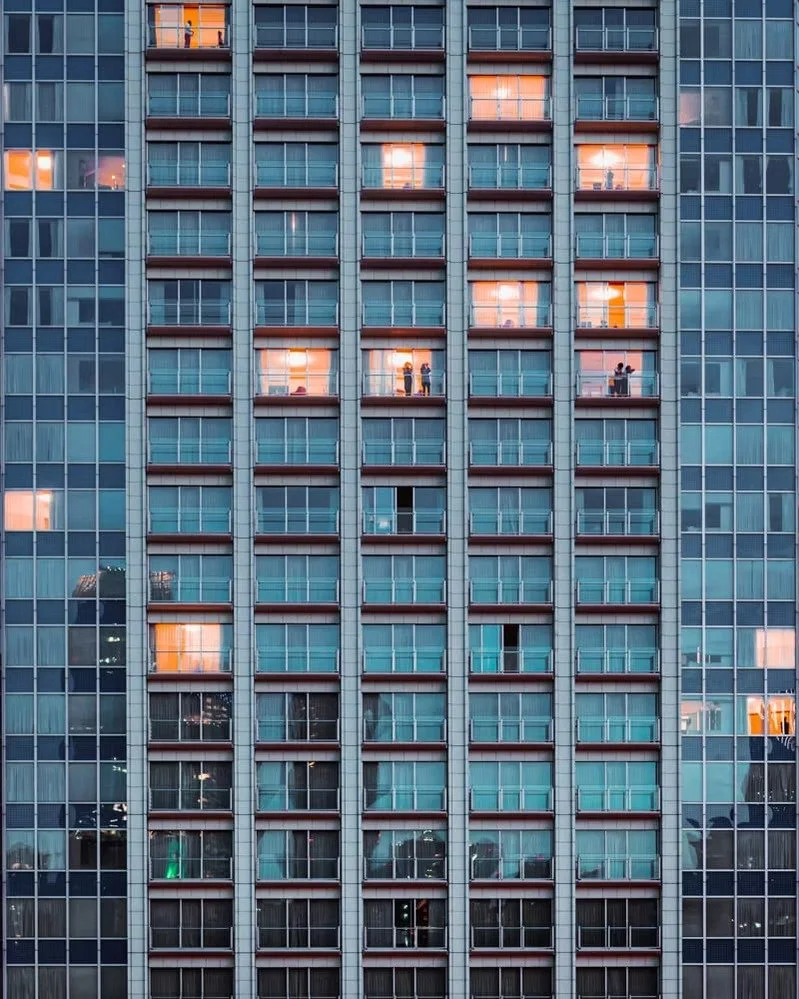 A tall office building with numerous windows, some illuminated, showing various people working or standing inside.