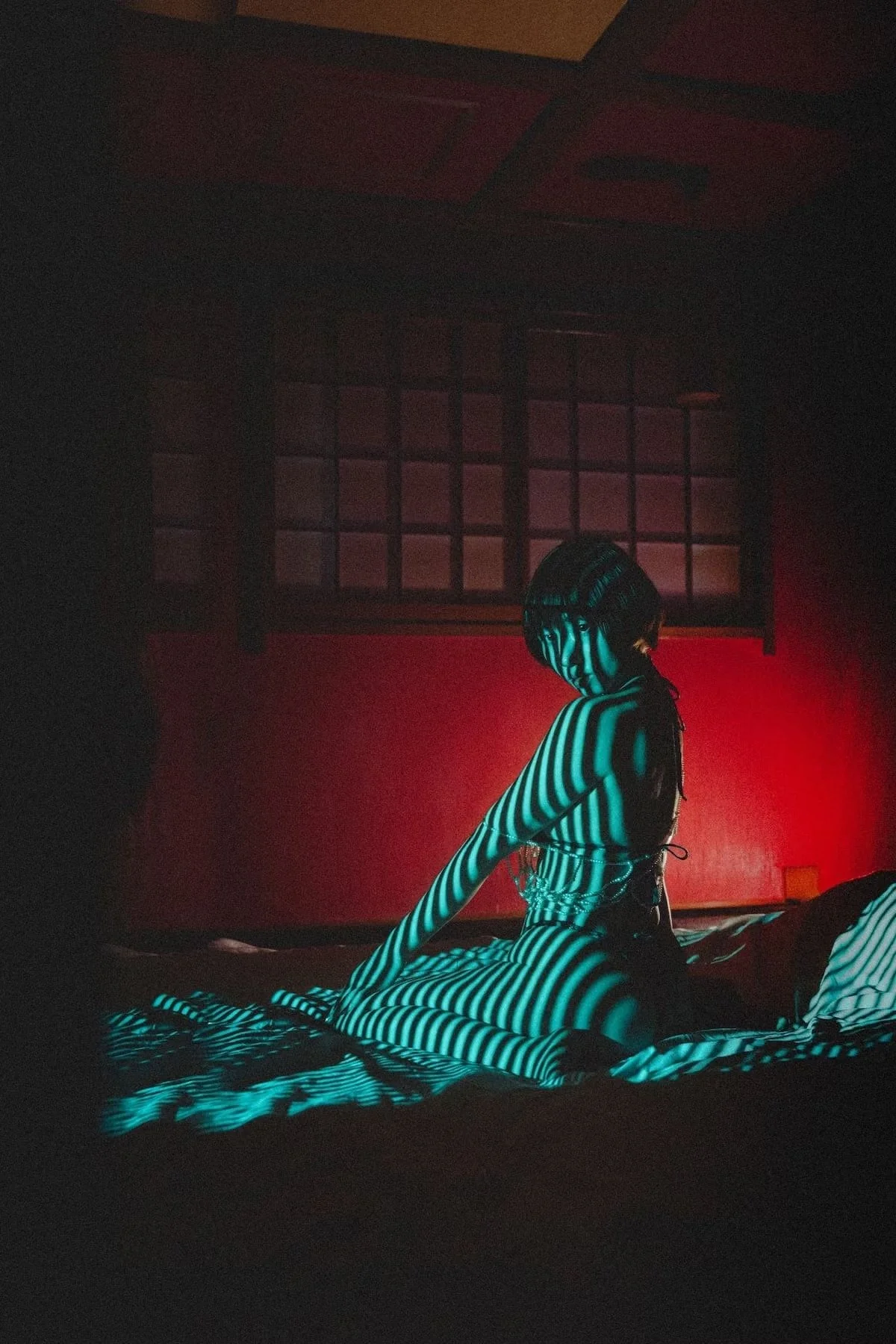 A person with striped clothing sitting on a bed in a dimly lit room with red and black walls, and a window in the background.