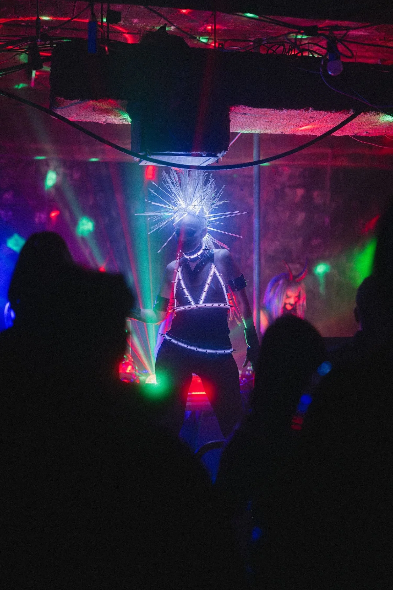 Bondage Ball 
---
Person wearing a headpiece and outfit with LED lights, performing in front of colorful laser lights and audience silhouettes in a dark setting.