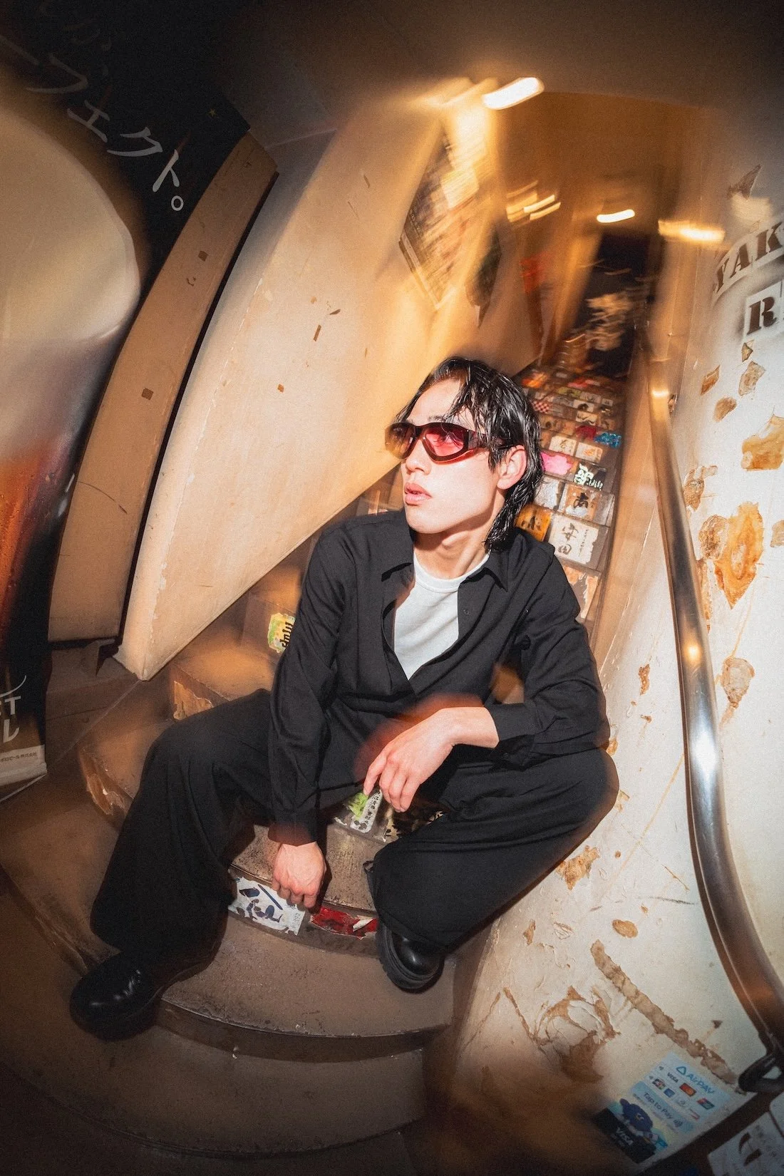 Vasuma Eyewear
---
A young man with black hair, wearing sunglasses, a black shirt, and black pants, sitting on a spiral staircase in a small, dimly lit space with decorative walls and posters.