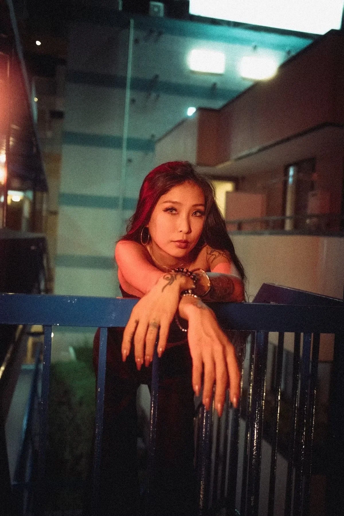 Portrait photography of a woman with dark hair and tattoos resting her arms on a blue railing in a dimly lit indoor space, looking at the camera with a contemplative expression.