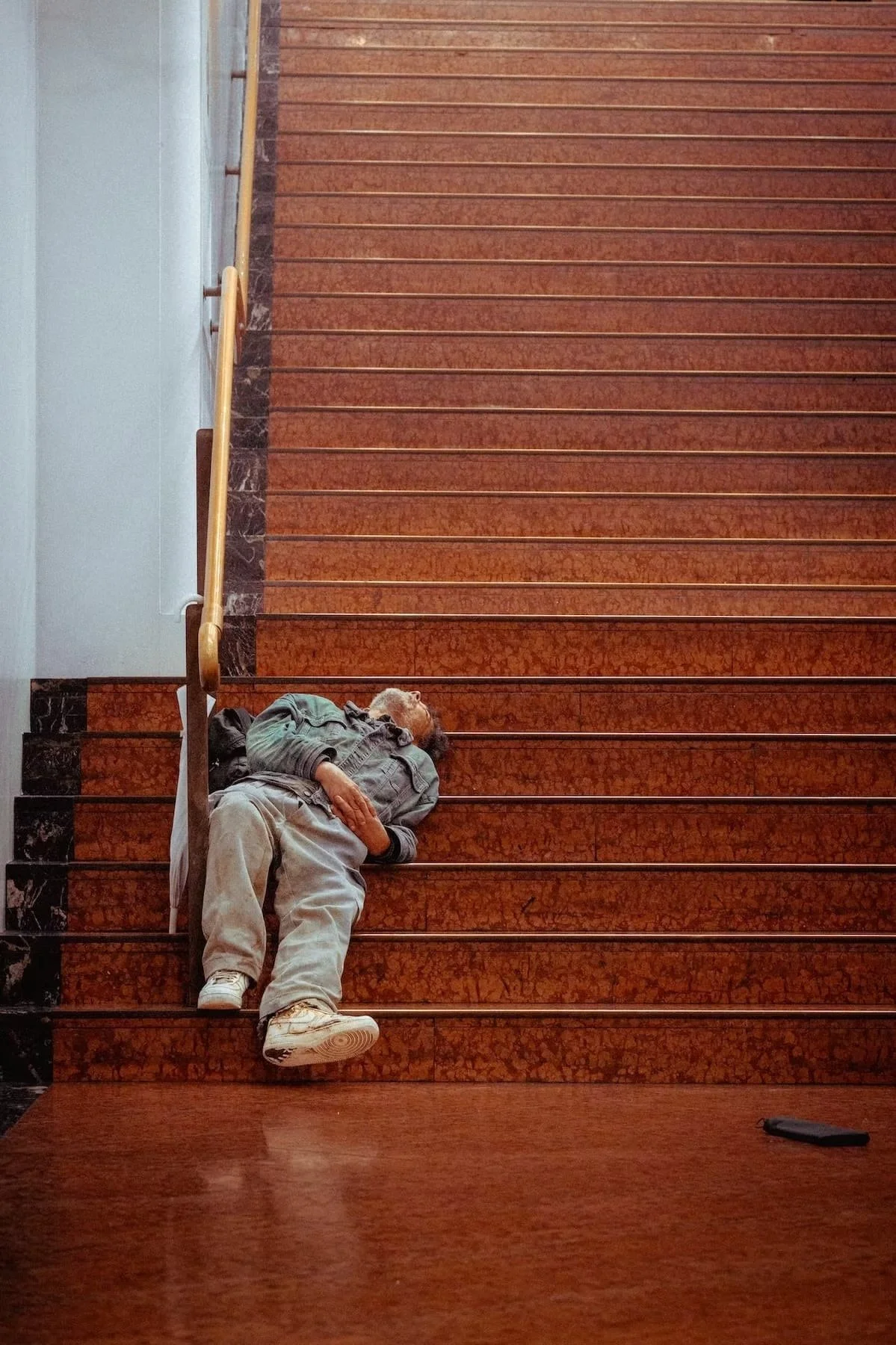 A person lying on the stairs of a building, resting on their side with eyes closed, wearing casual clothing including a denim jacket and pants, in an indoor setting with red marble stairs and a wooden handrail.