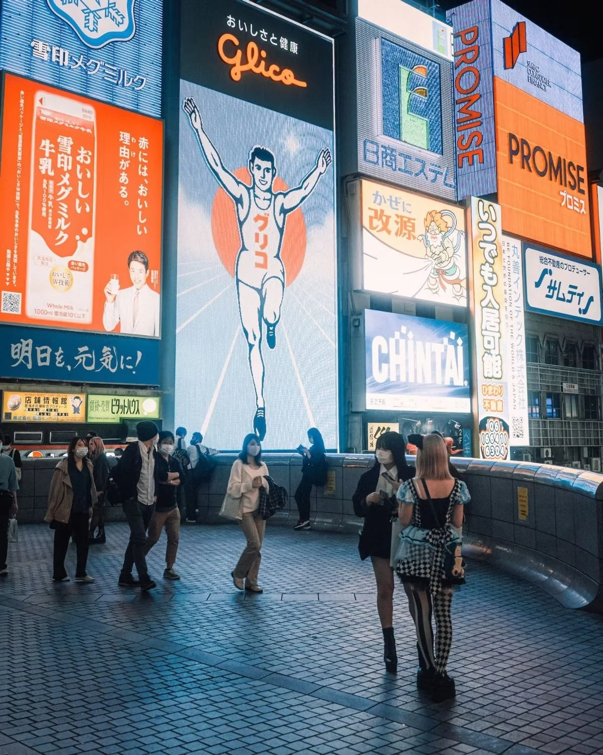 Nighttime scene in a busy city area with large illuminated billboards, including a running man illustration and various advertisements. Pedestrians wearing masks walk along the sidewalk. Dotonbory Glico running man.