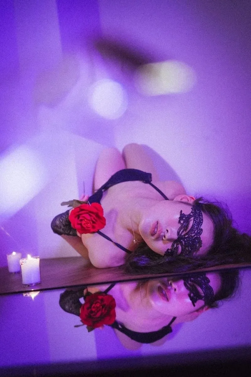 A woman in black lingerie with a red flower on her arm, wearing a lace mask, lying on the floor with candles nearby, and her reflection visible on a mirror.