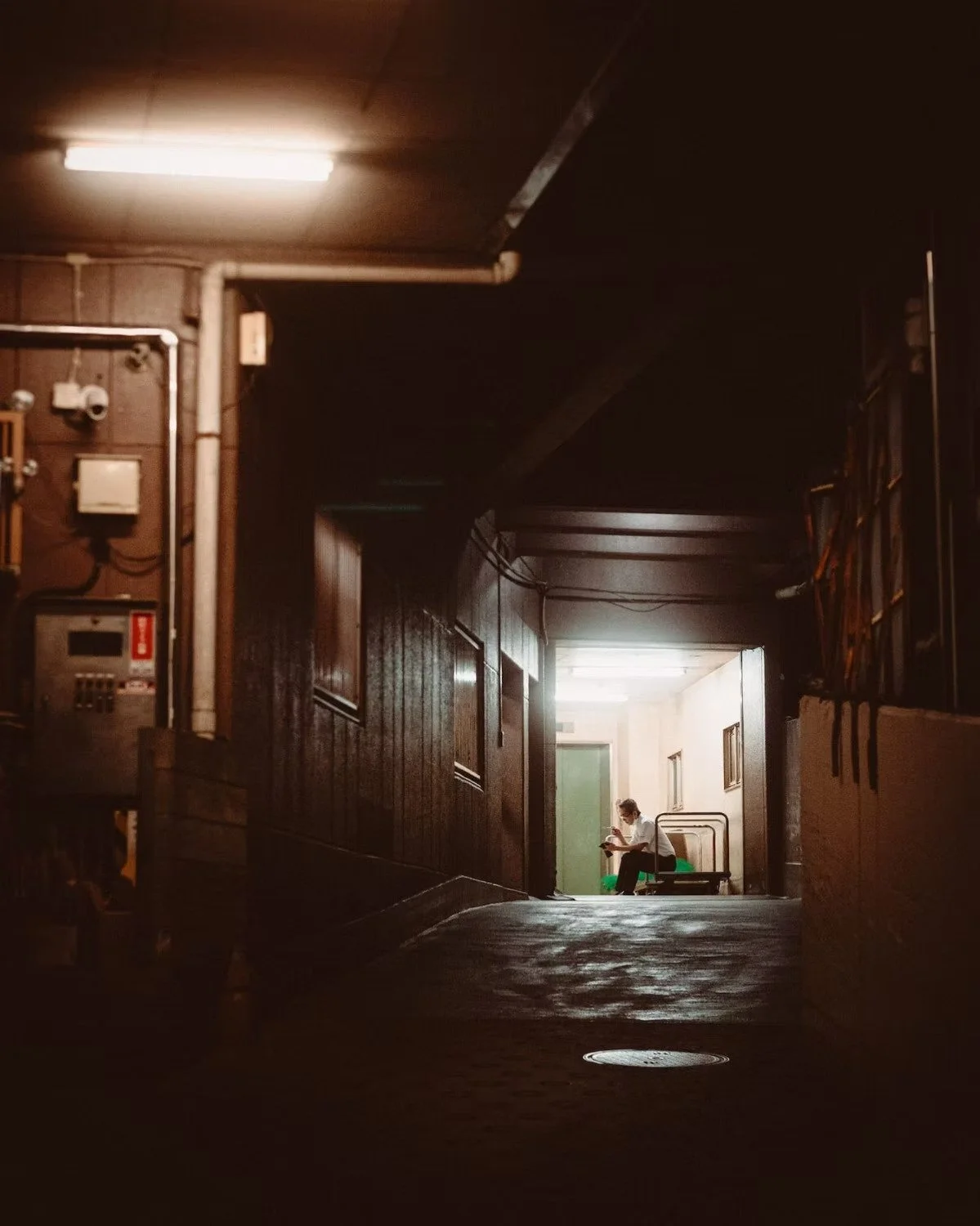 A person sitting on a bench reading a book or using a phone in a dimly lit alleyway with a bright exit at the end in Osaka.