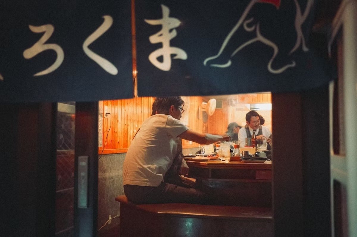 People dining at a Japanese restaurant, seen through a doorway. Two men are at a table, one serving food. Shimbashi Izakaya at night.