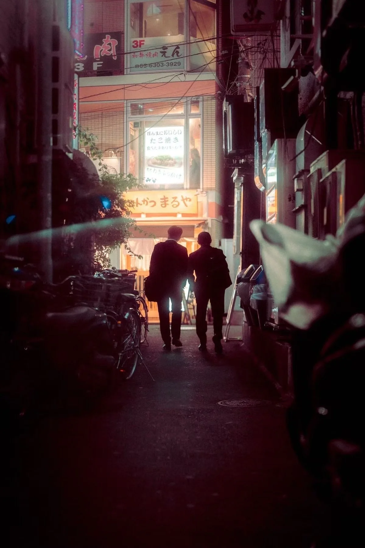 Two Japanese salarymen in business suits walking through a narrow alleyway at night in an urban area, lined with bicycles and illuminated shop signs in Japanese. Shot on the Tokyo Afterhours night street photography workshop by Tokyo street photograp