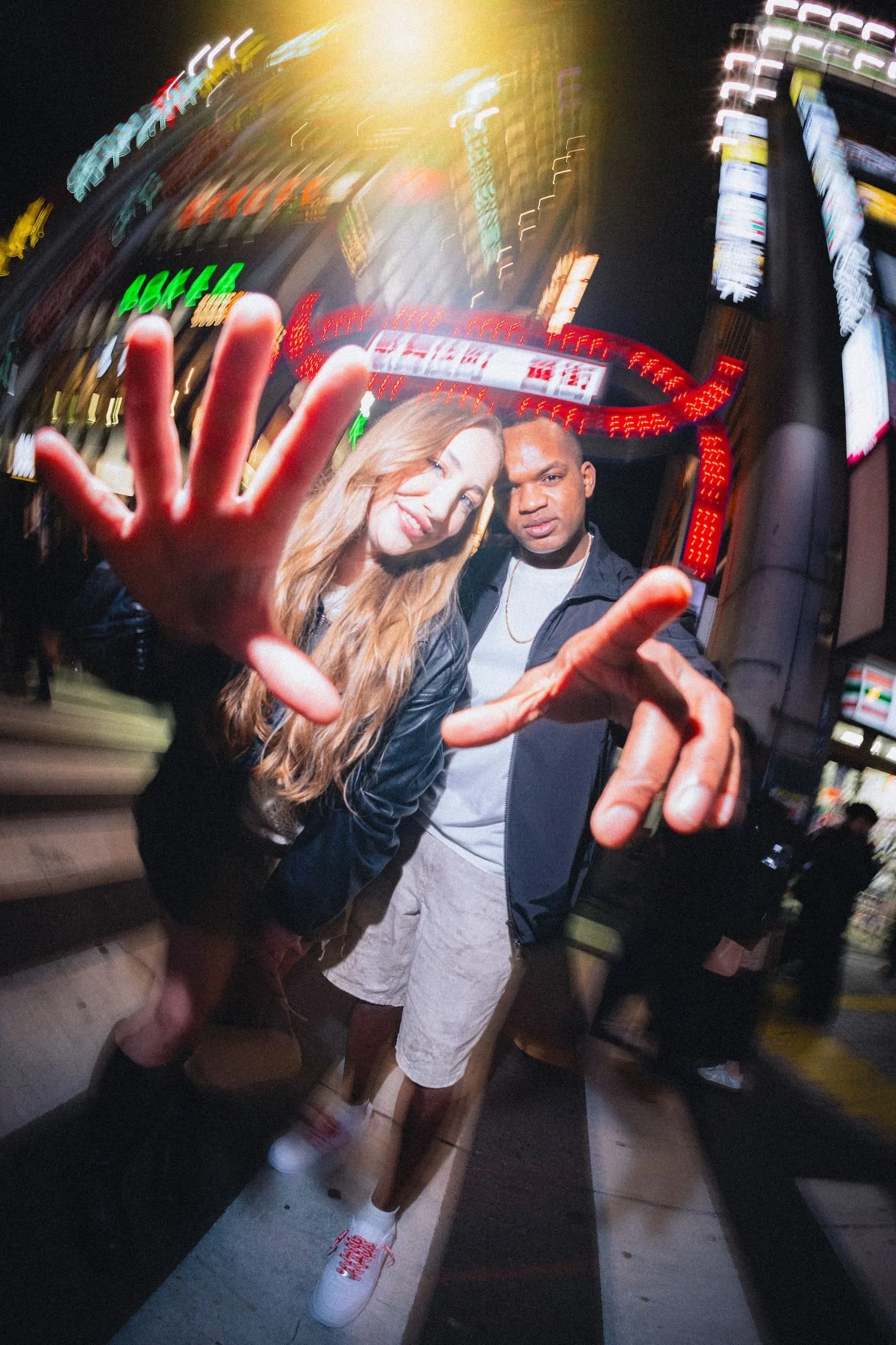 Two friends, one woman and one man, are posing playfully in front of a cityscape with bright neon signs and lights. The woman has long wavy hair and is smiling while winking, and the man is serious, wearing a chain necklace. They are making hand gestures, with the woman showing her palm towards the camera. The scene is lively and vibrant, indicating a busy urban nightlife.