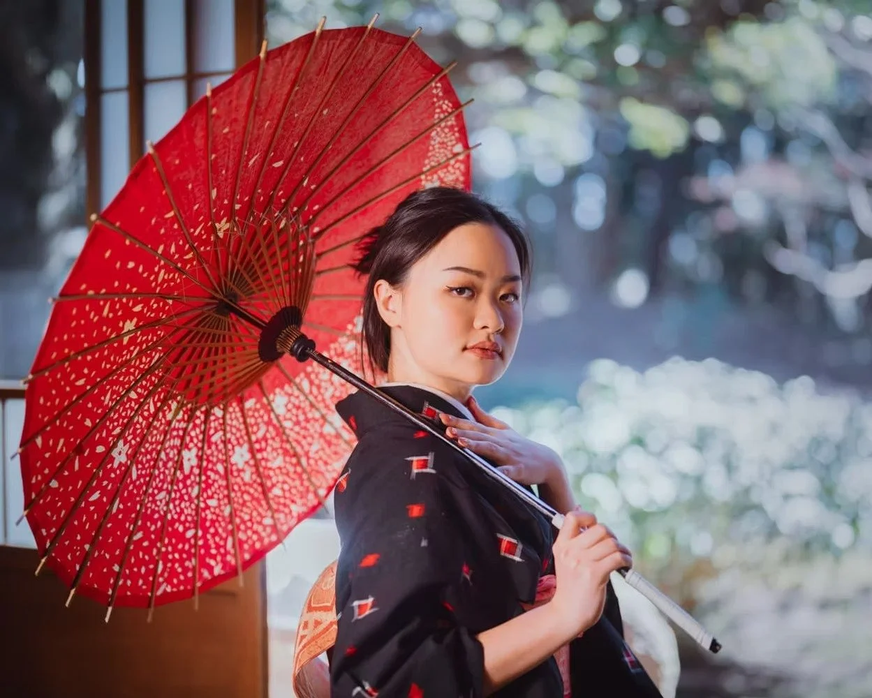 A woman wearing a black yukata holding a red paper umbrella, standing by a window with a blurred outdoor background.