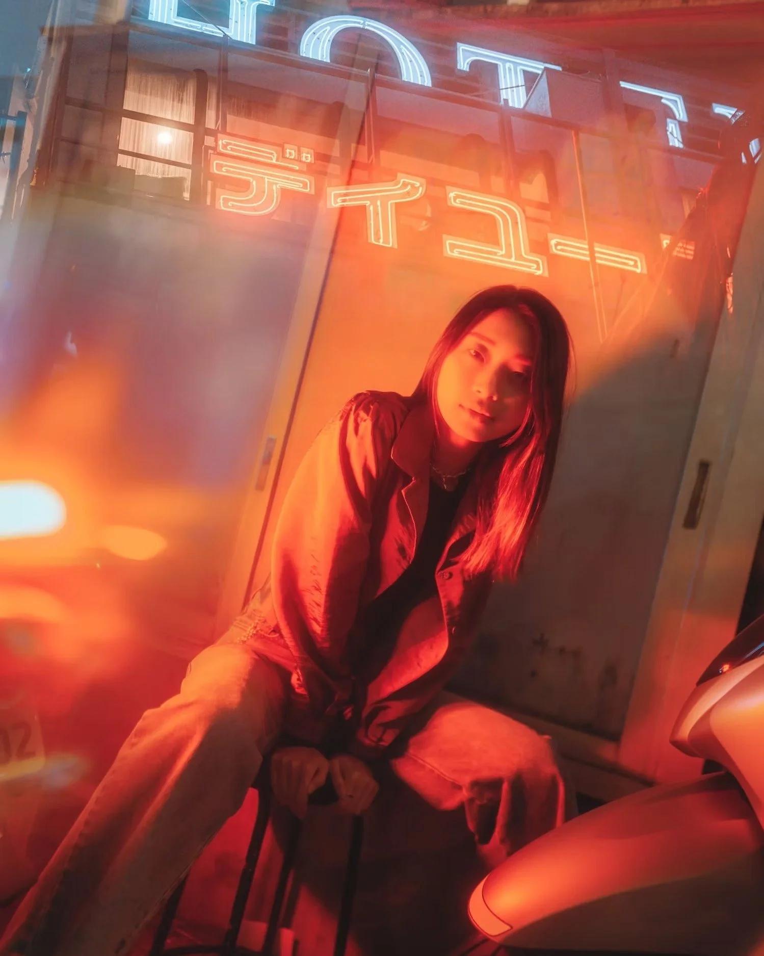 A young woman with long hair sitting inside a Tesla vehicle with red neon lighting and a visible Tesla sign on the building outside.