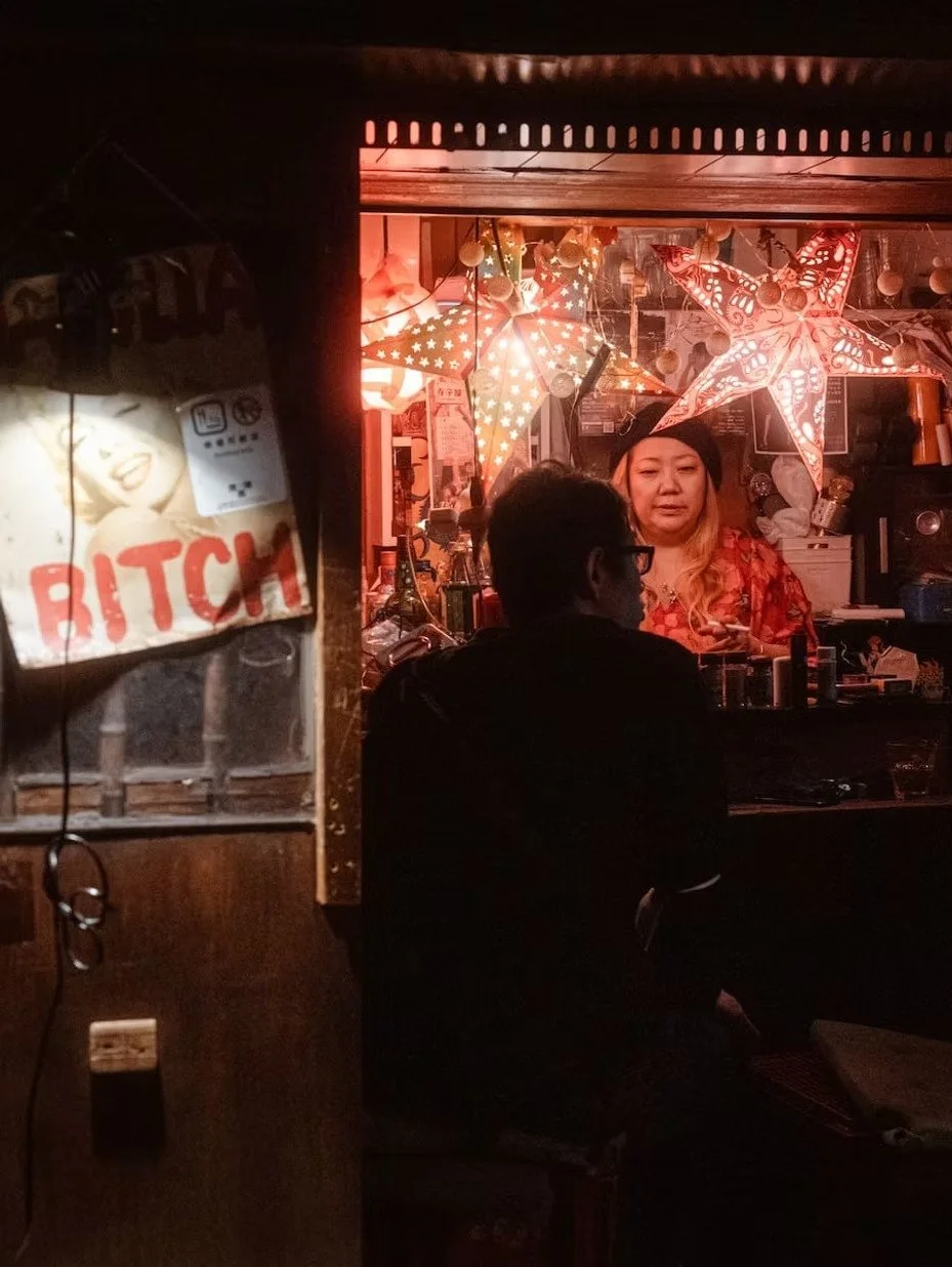 A dimly lit scene inside a bar with a woman behind the counter decorated with star-shaped and star-patterned lanterns. Two people are sitting on the other side of the counter, one with dark hair and glasses facing the woman, and another with a hat. A sign on the left side says 'BITCH' with an image of a woman smiling.