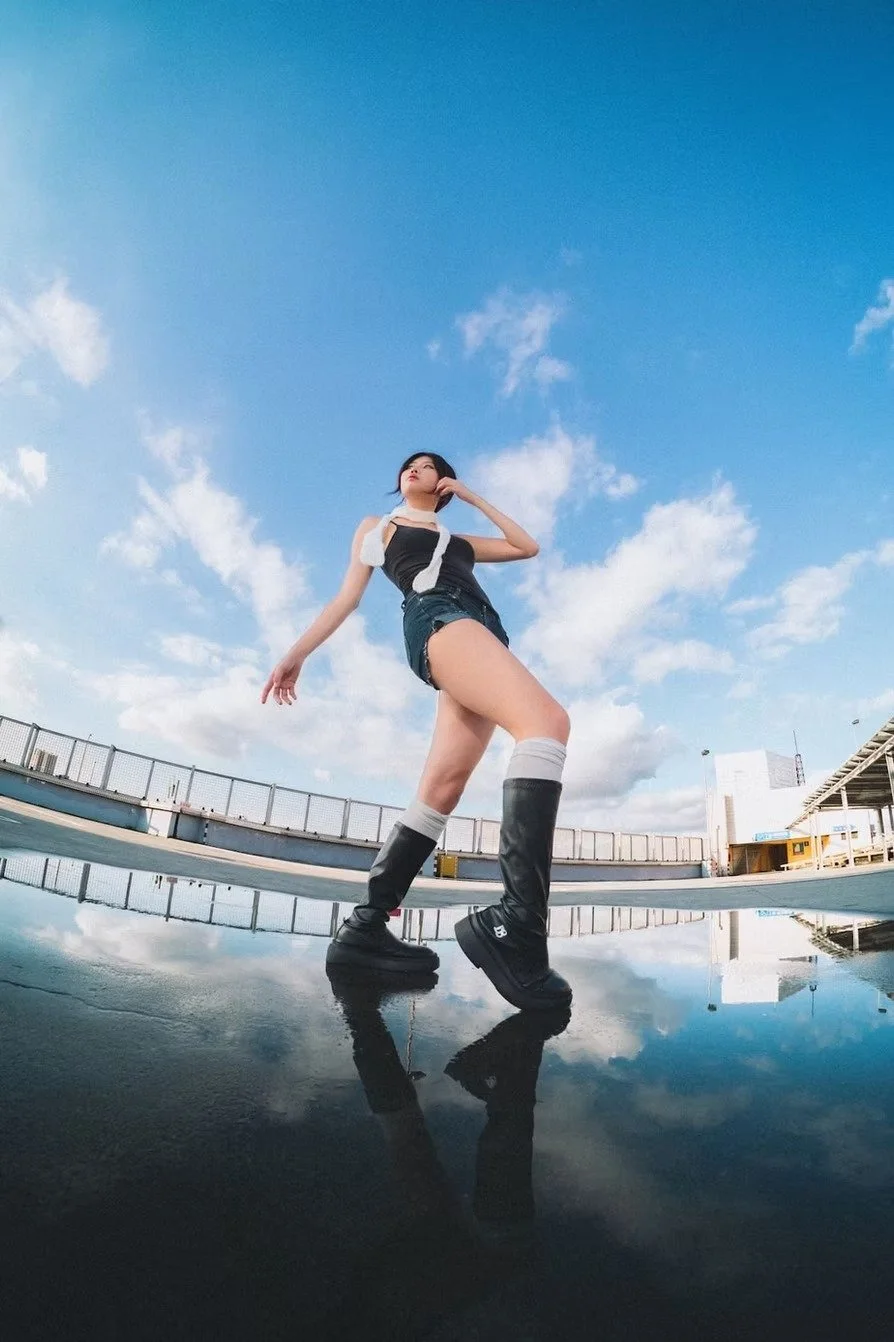 A young woman standing on a reflective wet surface outdoors, with a blue sky and clouds overhead, posing confidently with one hand touching her face and the other hanging by her side, wearing black boots, denim shorts, and a black and white outfit. F