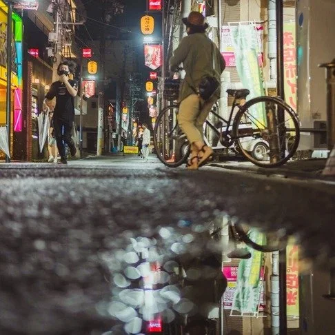 A cyclist pauses on a wet Tokyo street, neon signs reflecting in puddles, while a masked pedestrian walks past illuminated lanterns.