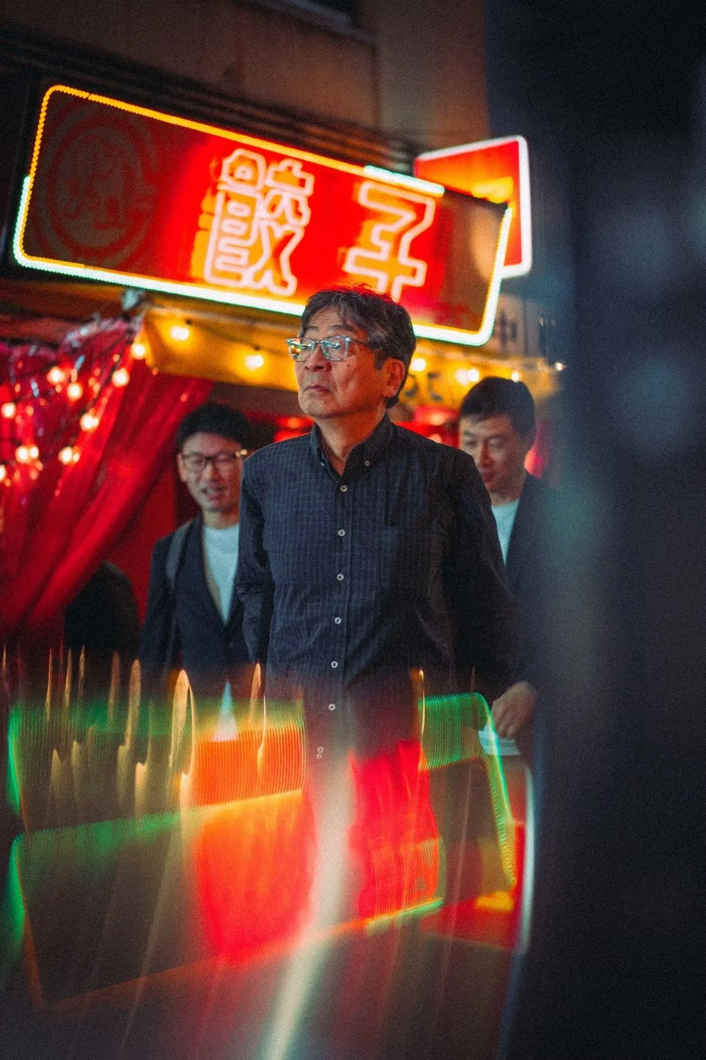 Nighttime scene in an urban area with a brightly lit neon sign in Japanese kanji, three men walking past the sign, colorful reflections on the wet surface, and a dark sky.