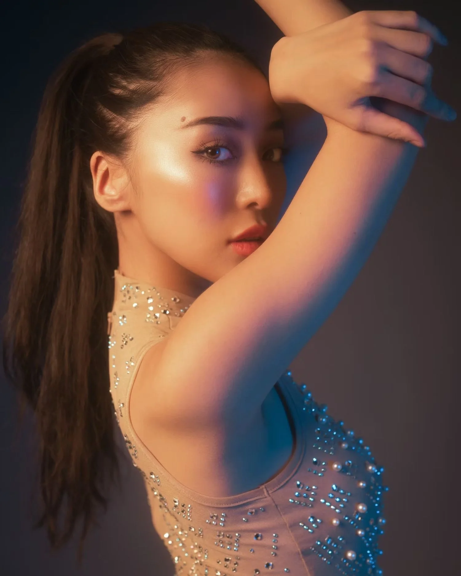A young woman with long dark hair and makeup, wearing a sparkly beige dress with rhinestones and pearls, with her arm raised in front of her face, looking at the camera with a confident expression, against a dark background.