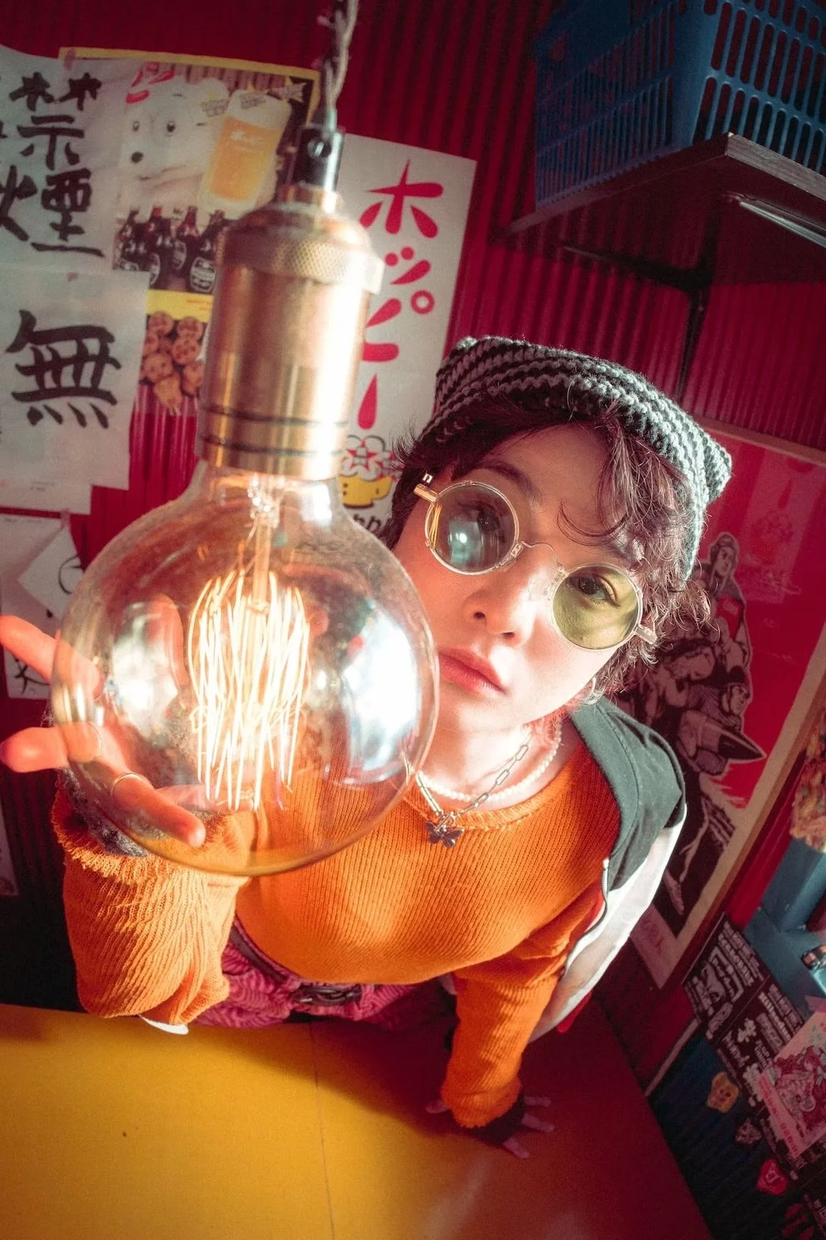 Wide angle portrait photography of a young person with short, curly hair wearing round glasses, a gray and black striped beanie, and an orange sweater, holding a large illuminated light bulb up to the camera inside a colorful establishment with Japan