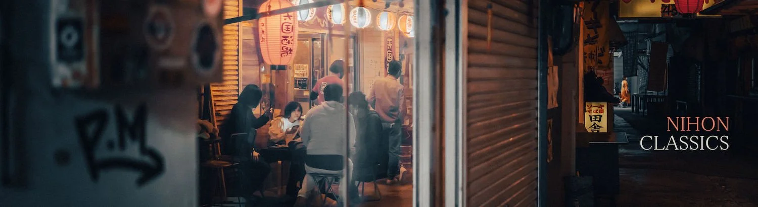 View of a Japanese restaurant at night through a glass door, with several people dining inside, illuminated by warm lighting, and Japanese signage outside.