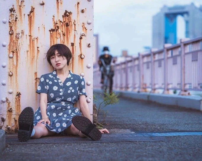 A girl sitting on a road with rusted walls and a distant cyclist, creating a unique composition in Osaka.