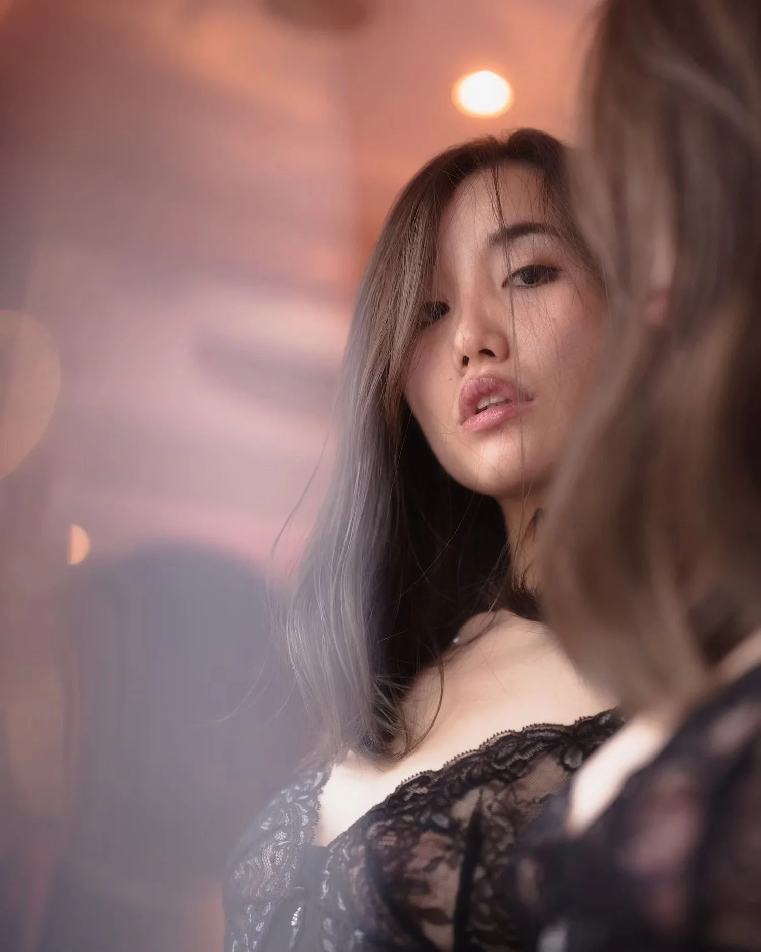 Close-up of a young woman with long, dark hair and makeup, wearing black lace clothing, with warm blurred lights in the background.