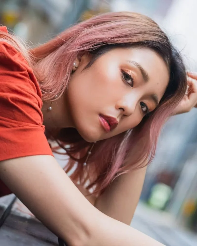 A young woman with pink hair, wearing a red top, posing outdoors with her head resting on her hand and gazing at the camera.